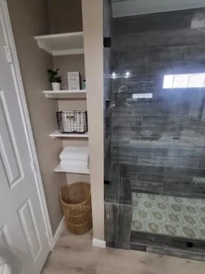 Frameless glass shower with dark tile, custom built-in shelving with white shelves and woven basket storage
